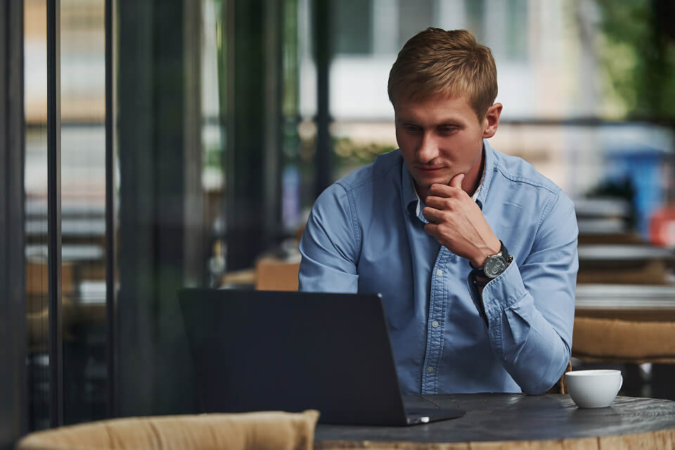A man with dirty blonde hair is resting his chin on his left hand while sitting at a round table in a cafe. He is typing with his right hand while he intently reads his favorite newsgroups on Newshosting.