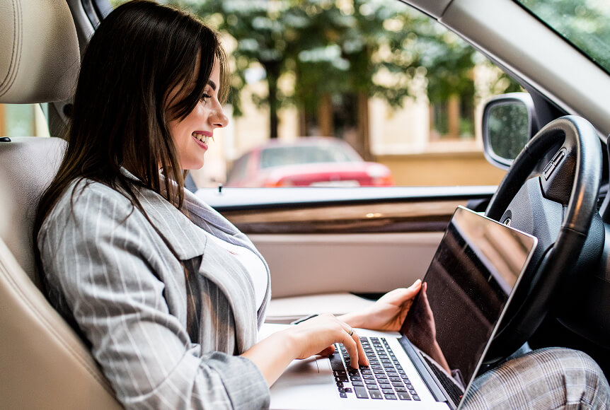 A dark-haired woman is sitting in her car, parked, and is browsing newsgroups through Newshosting on her laptop while sitting behind the steering wheel.