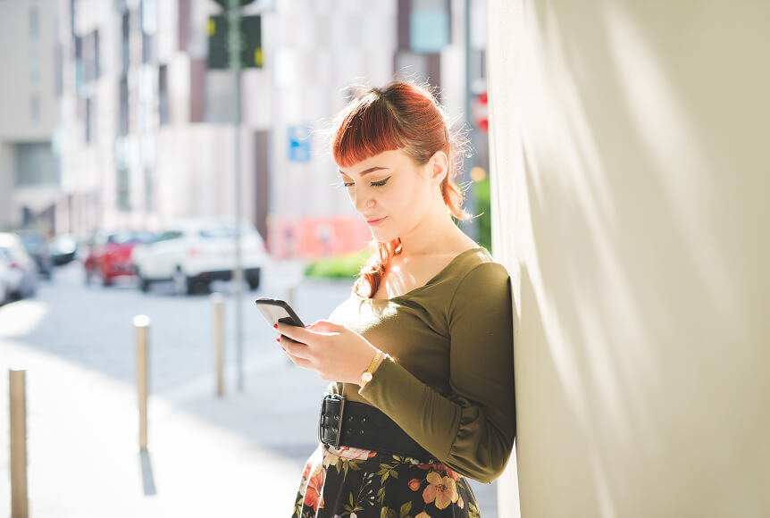 A redheaded woman in a deep green top and black, flowered skirt is leaning against a wall and smiling while she looks at her phone.