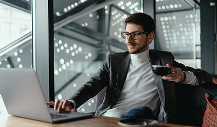 A man wearing a white turtleneck and sport coat is sitting at a conference table holding a black tea cup in his right hand while typing with his left hand on a laptop.